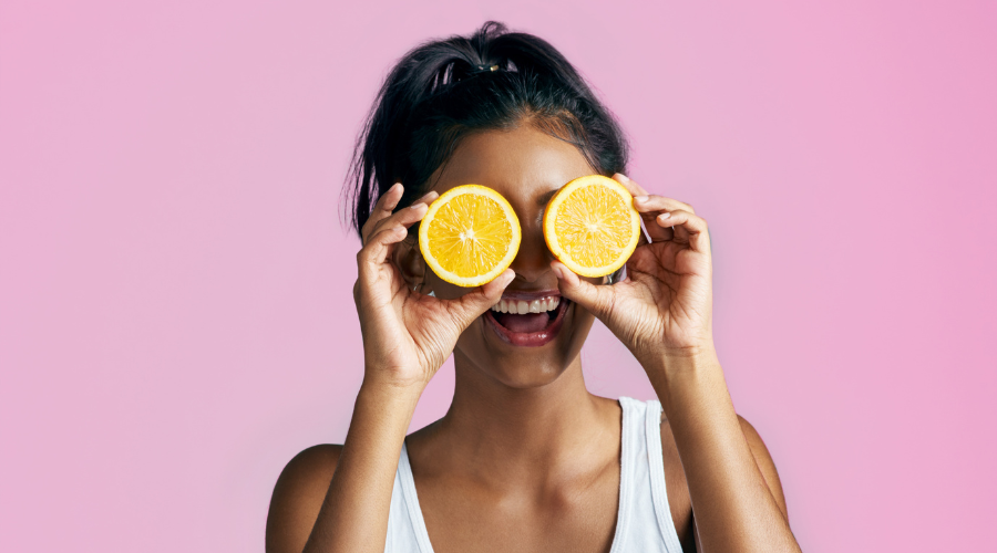 woman with brown hair holding two oranges in front of her eyes and smiling