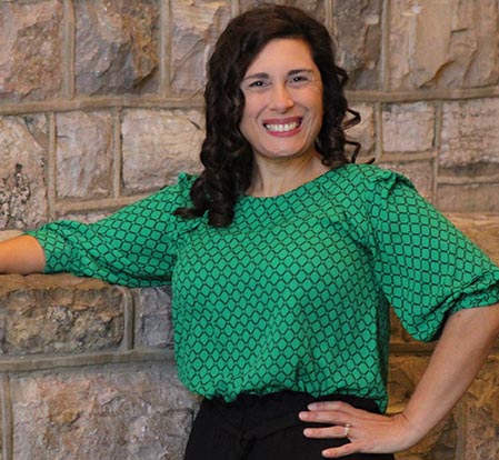 A smiling woman with dark hair wearing a green blouse stands in front of a stone brick wall